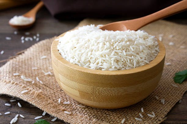 Close up shot of uncooked white Basmati rice in a wooden bowl with a wooden spoon.
Shot on a rustic wooden kitchen table with kitchen utensils in the background.